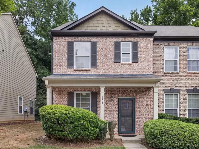 Brick facade townhome featuring a covered porch, dark shutters, and mature front landscaping