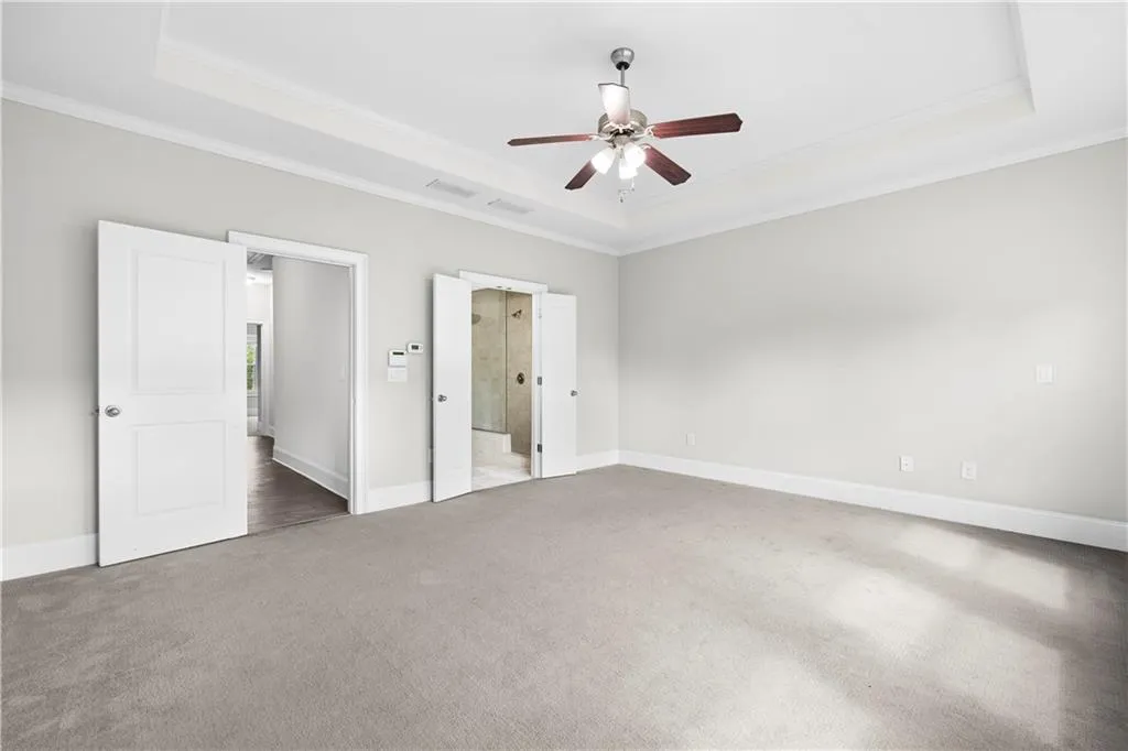 Unfurnished bedroom featuring a raised ceiling, carpet, crown molding, ceiling fan, and ensuite bath