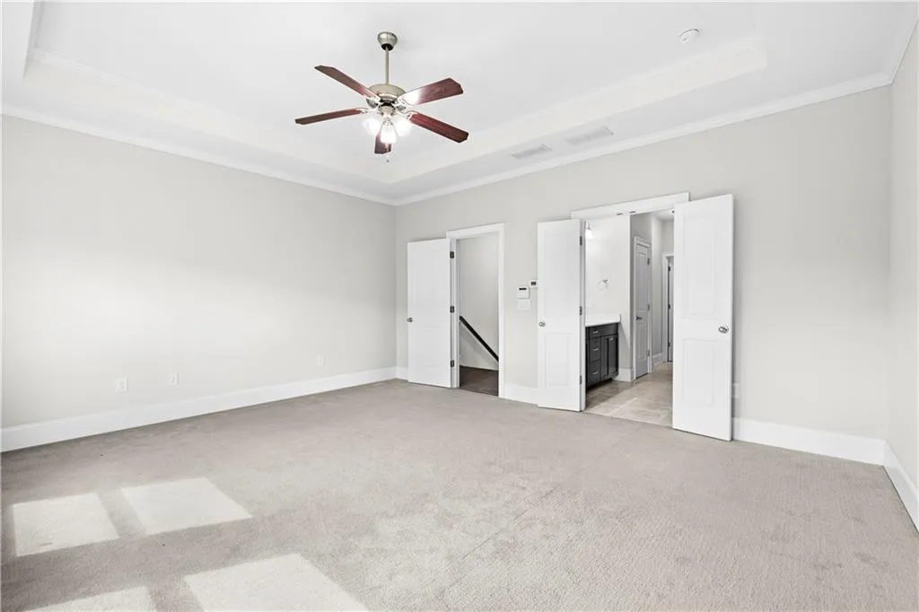 Unfurnished bedroom with a raised ceiling, light colored carpet, crown molding, ceiling fan, and ensuite bathroom