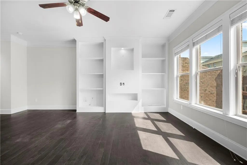 Unfurnished living room with built in features, ornamental molding, dark wood-style flooring, and ceiling fan