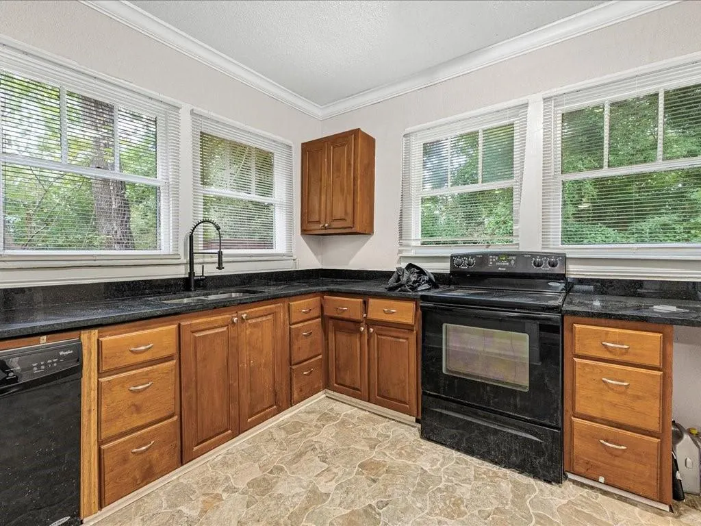 Kitchen featuring black appliances, brown cabinetry, light stone finish floors, dark stone counters, and crown molding