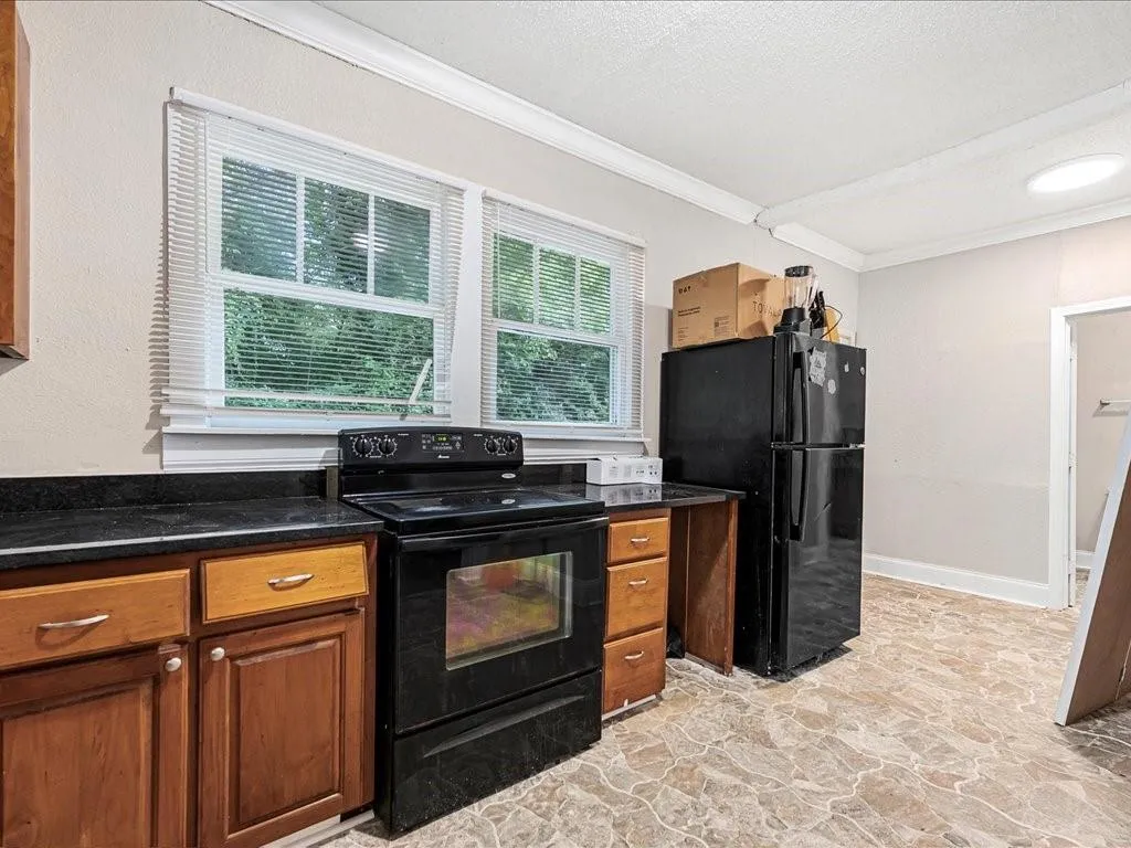 Kitchen featuring black appliances, light stone finish floors, crown molding, brown cabinets, and dark stone countertops