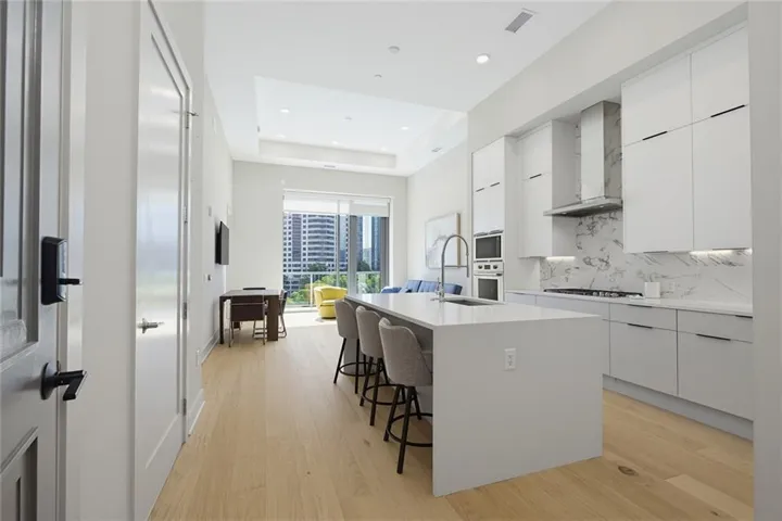 Modern kitchen featuring a waterfall island, integrated sink with gooseneck faucet, light wood-finish flooring, white flat-panel cabinetry, and a full-height backsplash