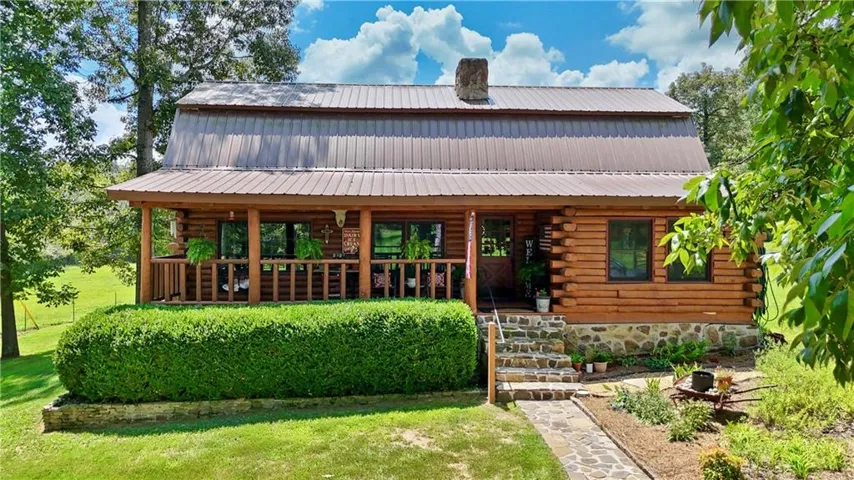 Charming log-cabin-style residence featuring a welcoming front porch with wood railings and multiple windows, metal roof, and a manicured landscape with mature hedging and established trees