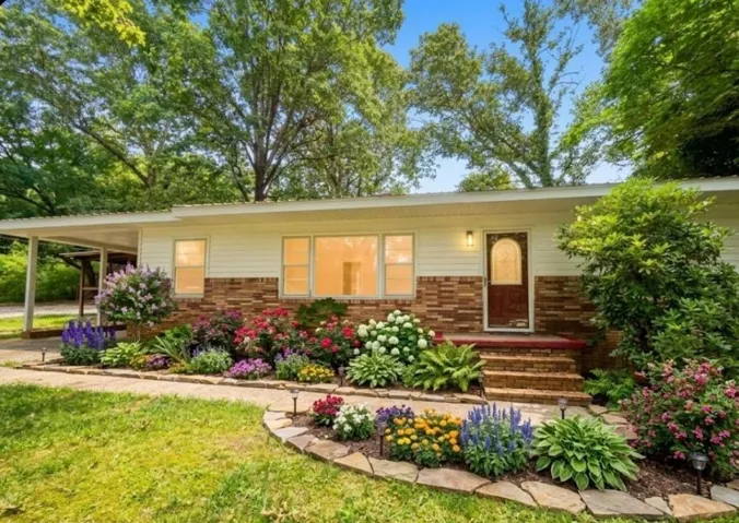 Brick and siding exterior with a covered carport, mature trees, a stone pathway, and extensive landscaping with various flowering plants