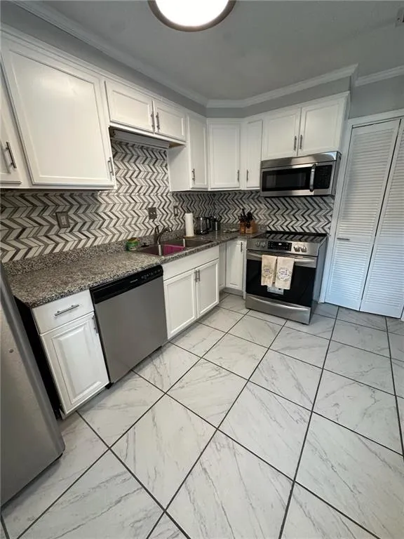Kitchen featuring crown molding, light marble finish flooring, decorative backsplash, appliances with stainless steel finishes, and white cabinetry