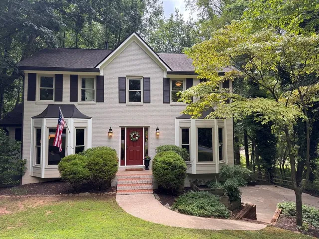 Brick facade residence featuring a dark shingle roof, black window shutters, a red entry door, and a curved concrete walkway