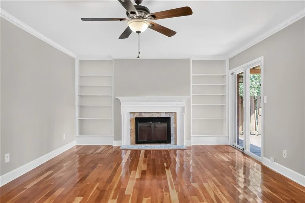 Hardwood flooring, fireplace with stone surround and white mantel, built-in shelving, ceiling fan, and sliding glass door