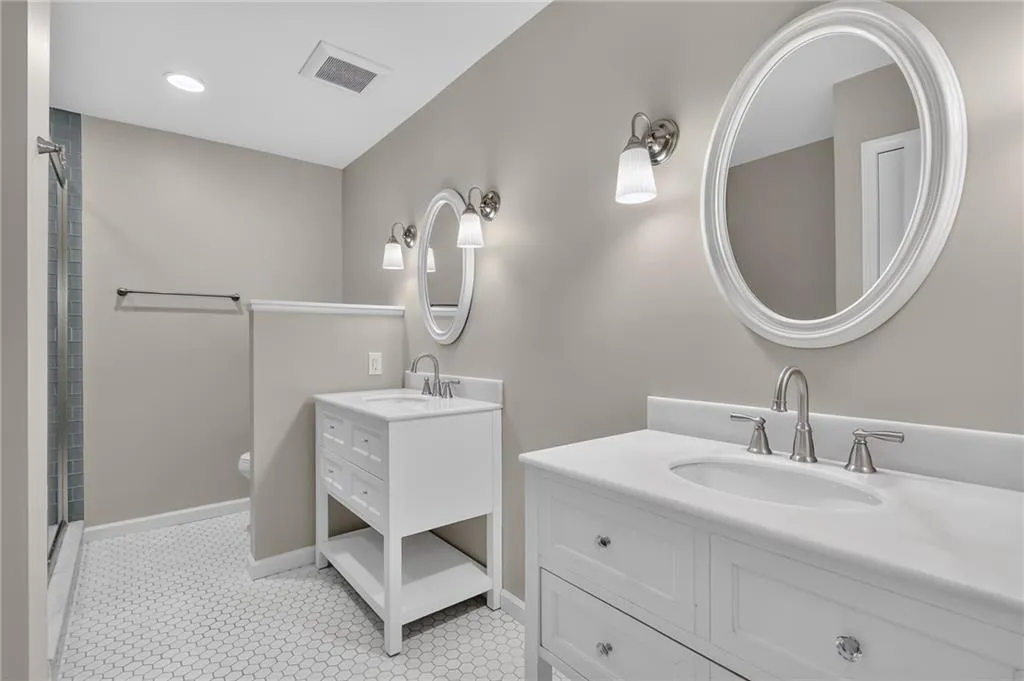 Dual white vanity sinks with coordinating oval mirrors, white hexagon tile flooring, and a tiled shower stall