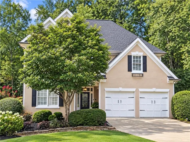 Hardcoat stucco exterior with a dark shingle roof, featuring white-framed windows with black shutters, a two-car garage with white doors, and a stone-edged planting bed