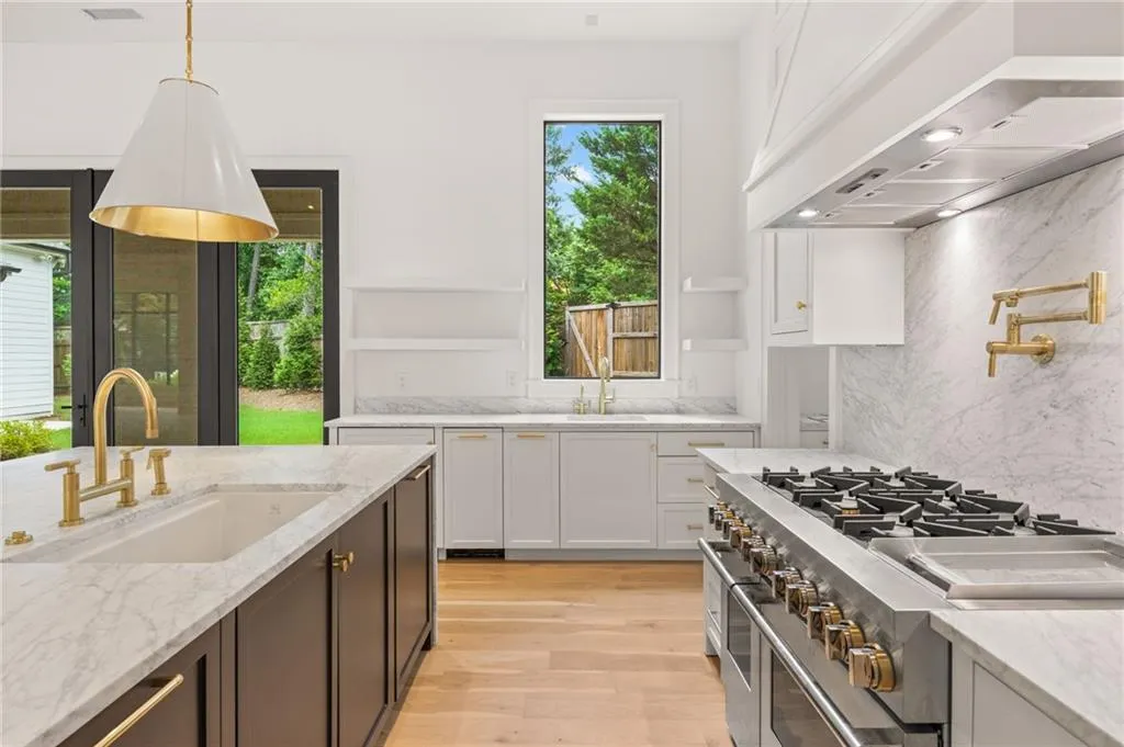 Spacious kitchen featuring a large island with an undermount sink and brass faucet, a professional-grade gas range with a pot filler, extensive white cabinetry, light wood-finish flooring, and white stone countertops and backsplash
