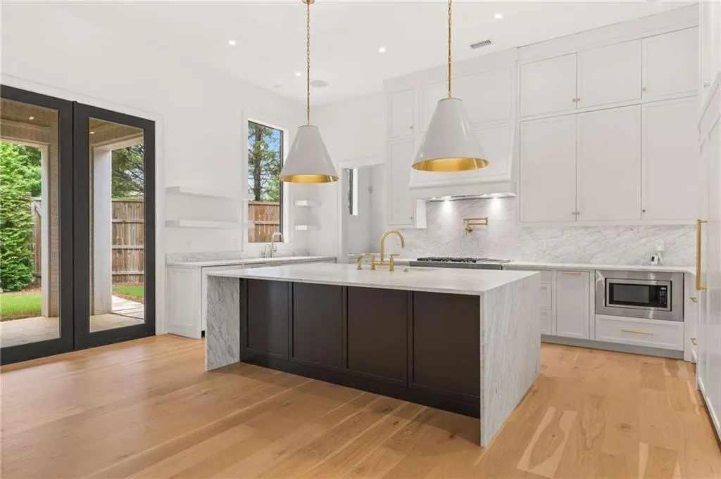 Contemporary kitchen featuring a large central island with waterfall countertop, white cabinetry with gold-tone hardware, integrated stainless steel microwave, and wood-finish flooring