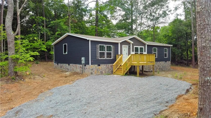 Dark gray siding exterior with stone accent skirting, a gravel driveway, and a wood deck