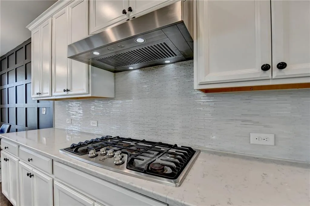 Kitchen with white cabinets, light stone countertops, under cabinet range hood, stainless steel gas cooktop, and backsplash