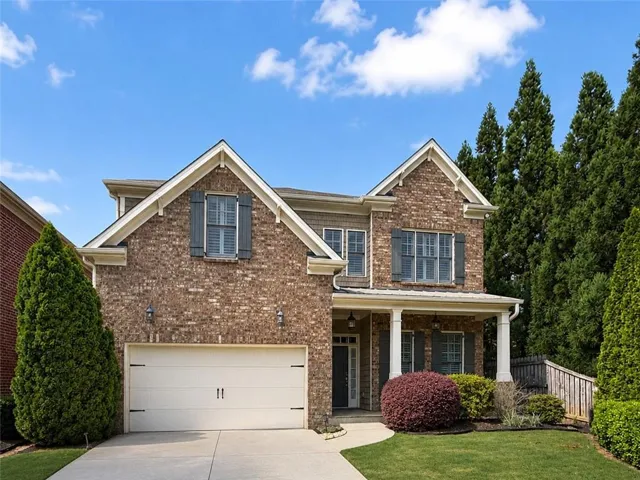 Brick facade residence featuring a two-car garage, covered porch with white columns, gabled rooflines, and decorative window shutters