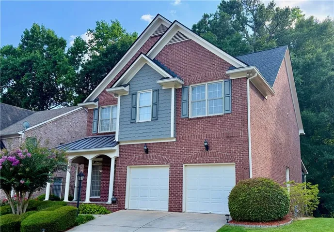 Brick facade residence featuring a two-car garage with white panel doors, a covered porch with white columns, and a dormer with blue shingle siding