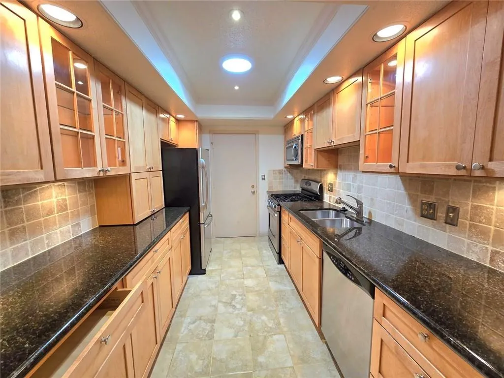Kitchen with glass insert cabinets, appliances with stainless steel finishes, tasteful backsplash, a raised ceiling, and dark stone counters