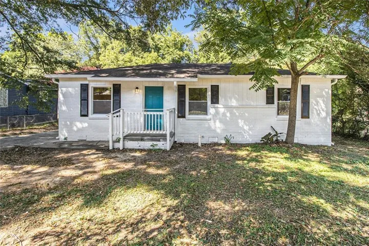 White brick exterior with a vibrant turquoise entry door, black louvered shutters, a front porch with white railing, and mature trees providing natural shade
