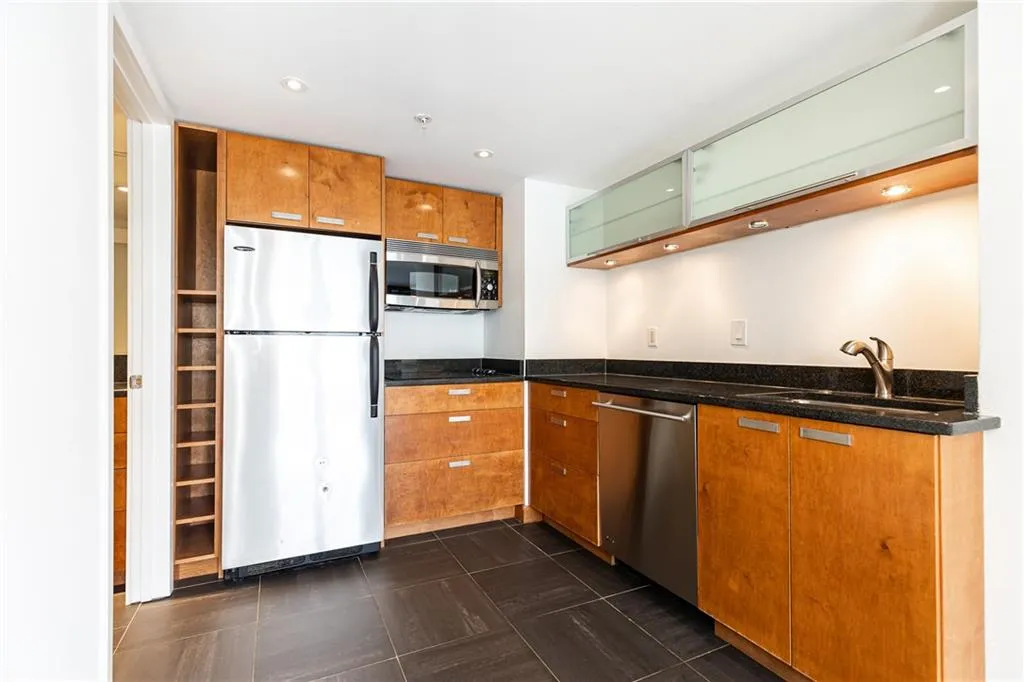 Kitchen with stainless steel appliances, dark stone counters, glass insert cabinets, brown cabinets, and recessed lighting