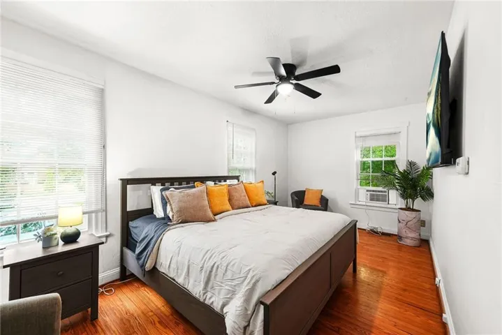 Bedroom featuring wood flooring, white walls, two windows with blinds, a ceiling fan with integrated lighting, and a wall-mounted television