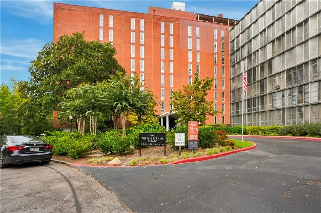 Red brick exterior, ample green landscaping, paved driveway, adjacent building with grid-patterned facade, flagpole