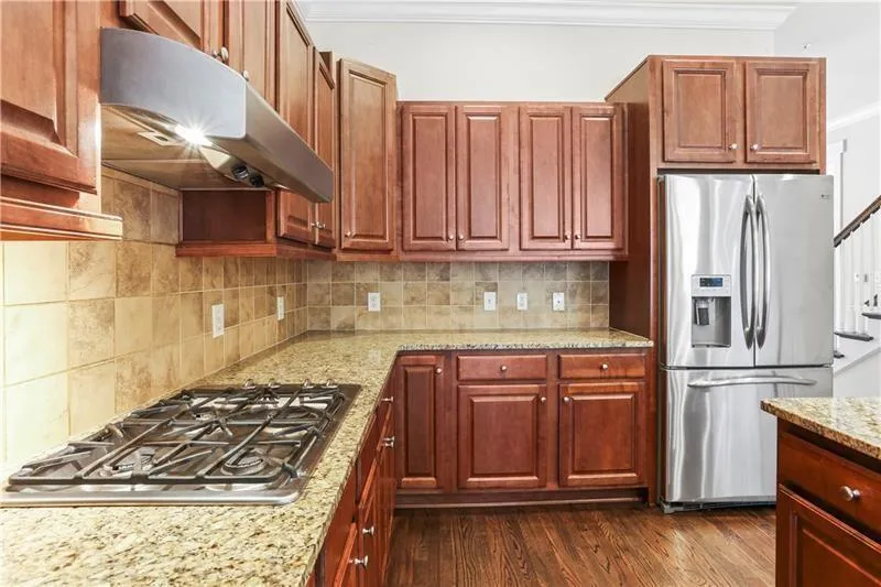 Kitchen with stainless steel appliances, under cabinet range hood, ornamental molding, tasteful backsplash, and light stone countertops