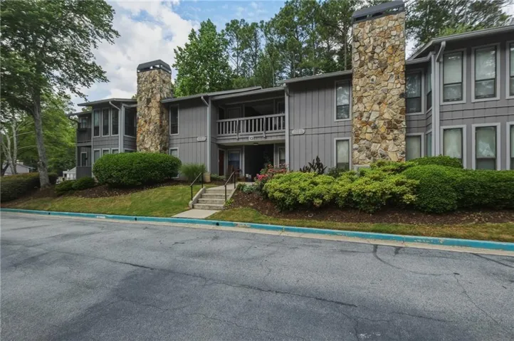 Exterior featuring two stone-clad chimneys, multiple windows, a second-story balcony, and mature landscaping