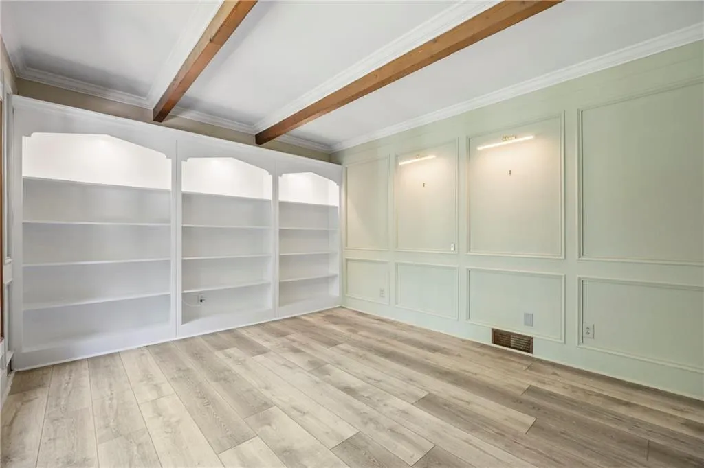 Empty room with light wood-style floors, crown molding, beamed ceiling, and a decorative wall