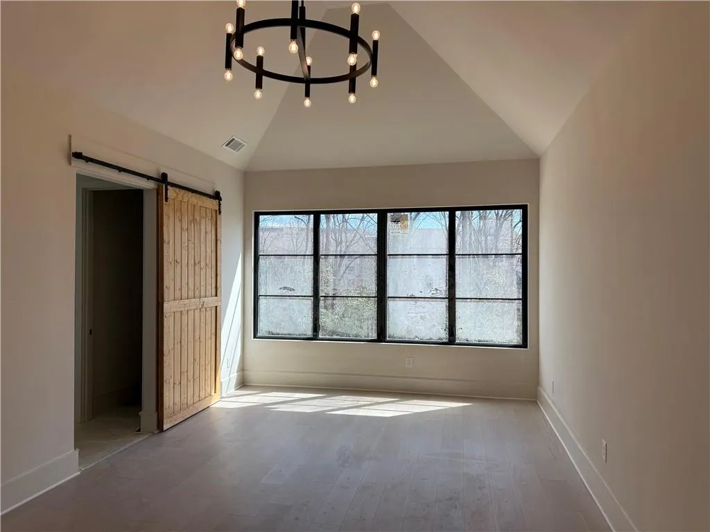 Unfurnished bedroom with a barn door, light wood-style floors, hanging lights, multiple windows, and lofted ceiling