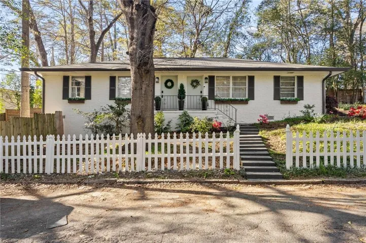 White brick exterior with dark shutters, front porch with black railing, concrete steps, and a white picket fence