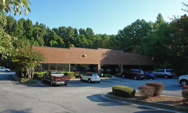 Brick exterior building with expansive windows, surrounded by mature trees, and an asphalt parking area with landscaped islands