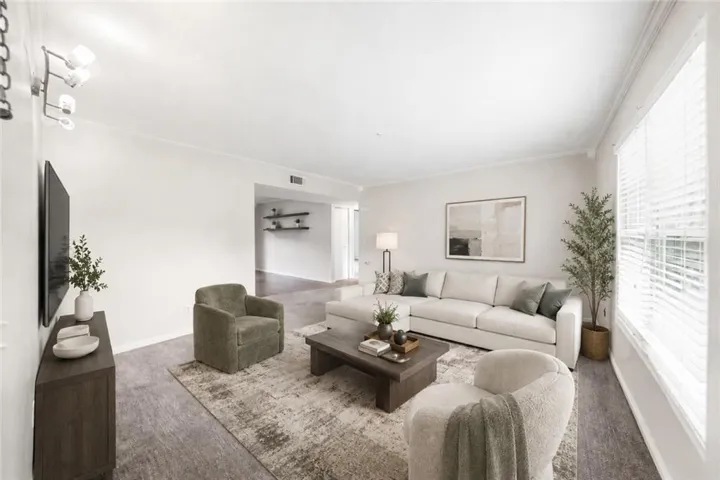 Spacious living area featuring light wood-finish flooring, white walls, crown molding, and a large window with blinds