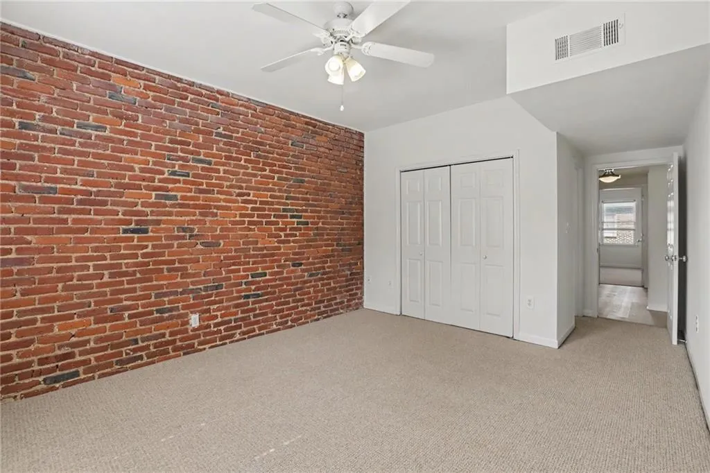 Exposed brick accent wall, neutral carpet flooring, ceiling fan with integrated lighting, bi-fold closet doors, and recessed air vent