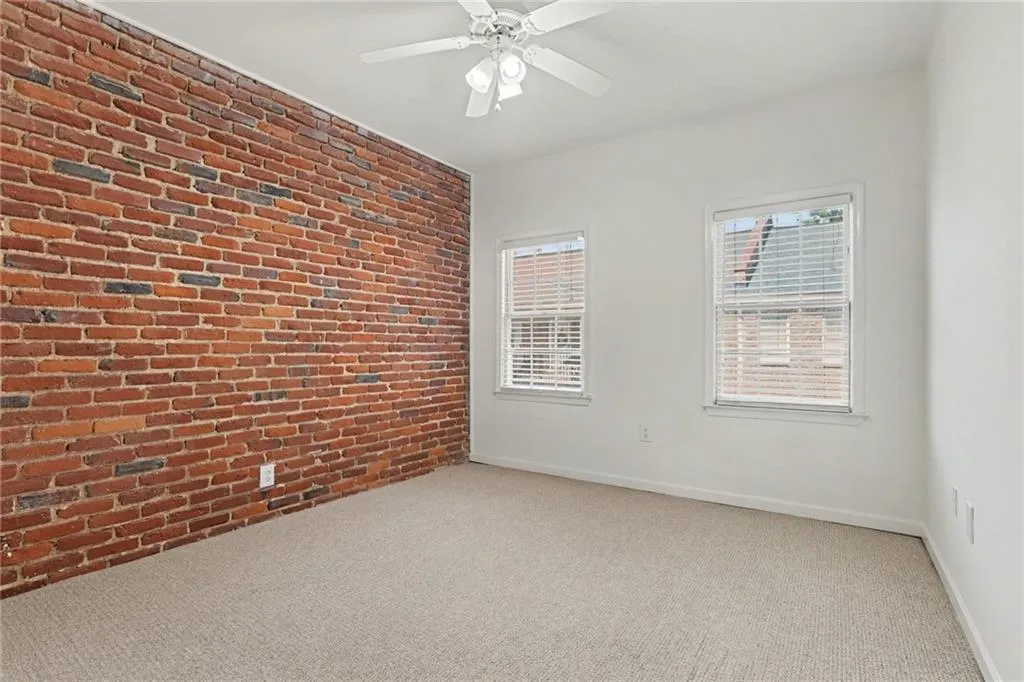 Exposed brick accent wall, neutral carpeting, two windows with blinds, and a ceiling fan