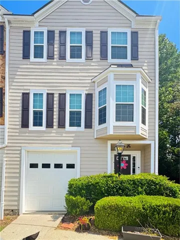 Three-story townhouse featuring a light-toned siding exterior, a prominent bay window, dark contrasting shutters, a single-car garage, and established front landscaping