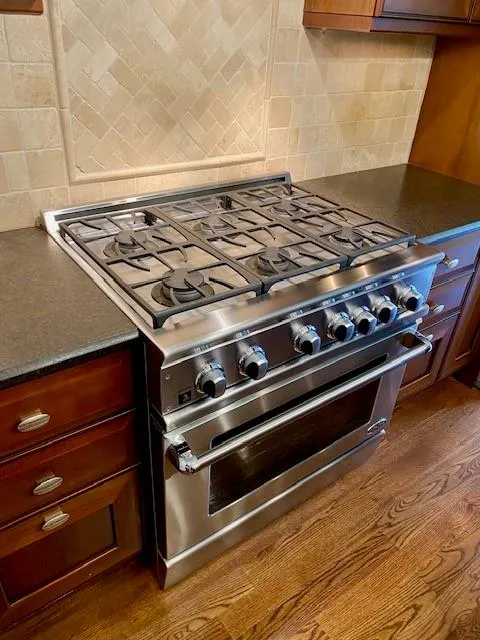 Kitchen view of high end range, decorative backsplash, dark stone counters, and dark wood finished floors