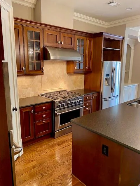 Kitchen with ornamental molding, stainless steel appliances, glass insert cabinets, under cabinet range hood, and decorative backsplash
