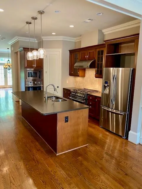 Kitchen featuring crown molding, dark countertops, appliances with stainless steel finishes, a center island with sink, and glass insert cabinets