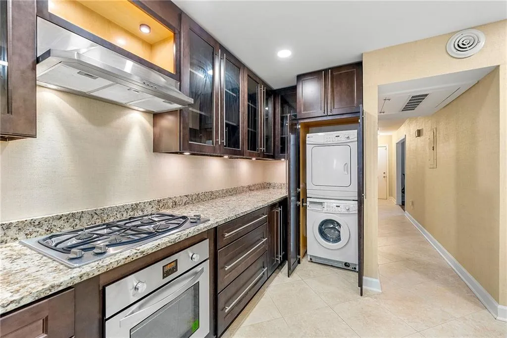Kitchen featuring light tile patterned floors, exhaust hood, dark brown cabinetry, stainless steel appliances, and stacked washer and clothes dryer