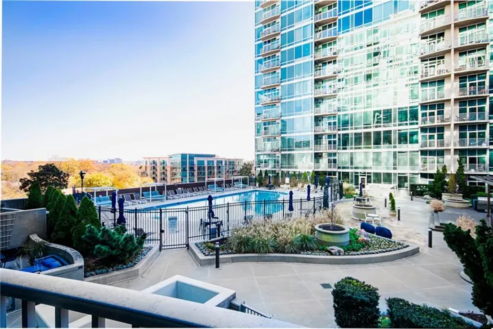 Expansive outdoor patio featuring a swimming pool, concrete hardscaping, mature landscaping, and an adjacent high-rise building with glass facades