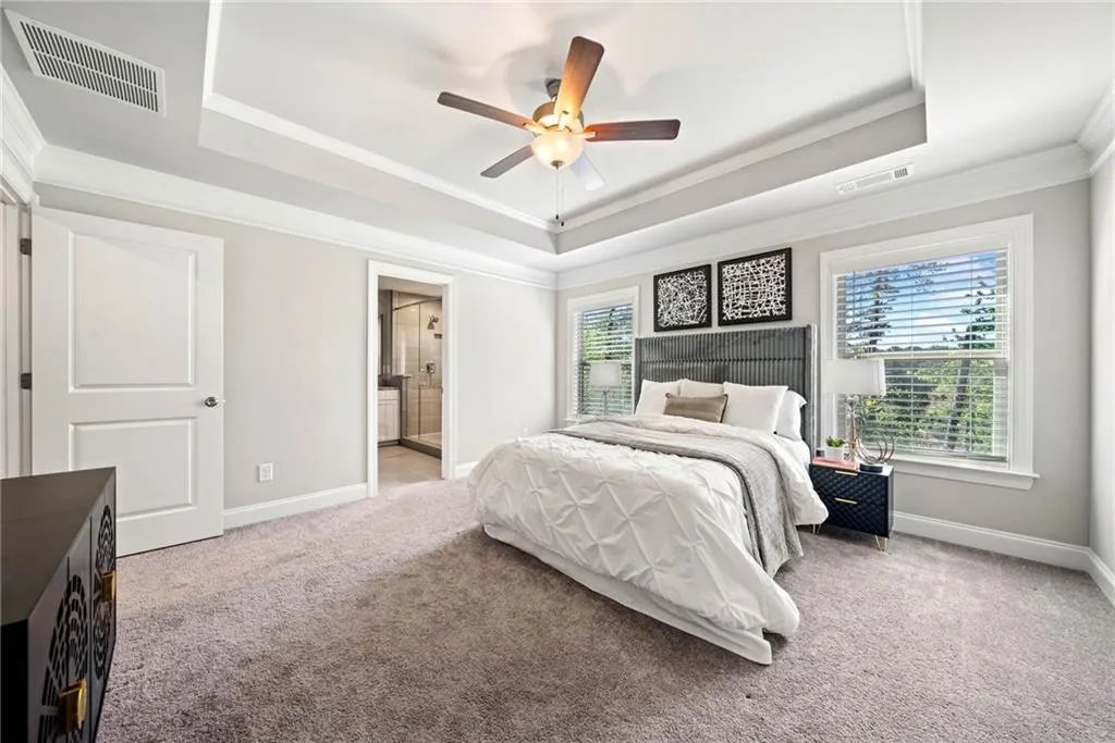 Bedroom featuring a raised ceiling, light colored carpet, ornamental molding, ensuite bath, and a ceiling fan