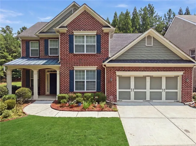Brick exterior featuring a covered front porch with columnar supports, a two-car garage with decorative doors, a concrete driveway, and a manicured lawn with established landscaping