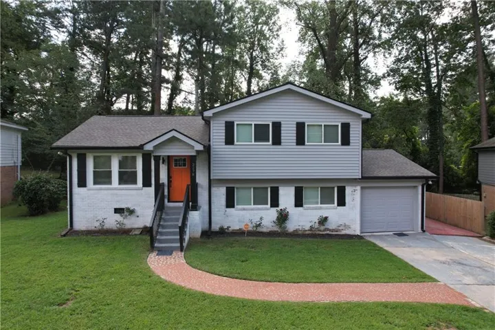 Split-level residence featuring gray siding and white brick, a vibrant orange front door, a single-car garage, a paved driveway, and a red brick pathway