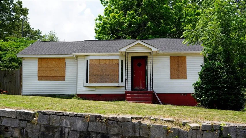 White siding exterior with a red front door, covered front porch, dark shingle roof, and a stone retaining wall