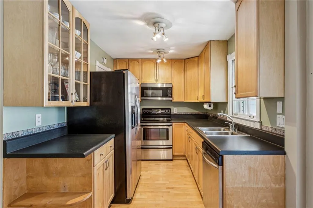 Kitchen featuring stainless steel appliances, dark countertops, light wood-style flooring, glass insert cabinets, and light brown cabinets