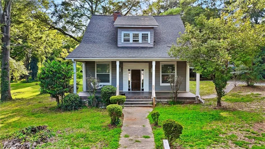 Gable roofline with dormer window, covered front porch with white columns, concrete walkway, and mature trees