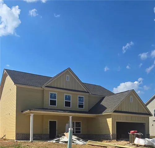 New construction residence featuring yellow siding, a dark shingle roof, a covered front porch with white columns, a two-car garage, and visible upper-level windows