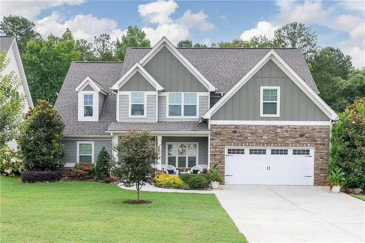 Craftsman-style home featuring a stone facade, shingled roof, board and batten siding accents, a two-car garage with decorative windows, and a landscaped front yard with mature plantings
