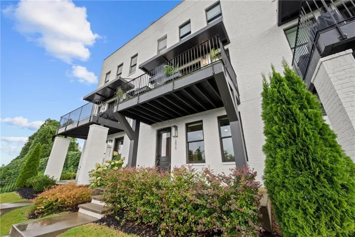 White brick exterior with black framed windows, multiple balconies with black railings, and a tiered landscaped entrance featuring shrubs and greenery
