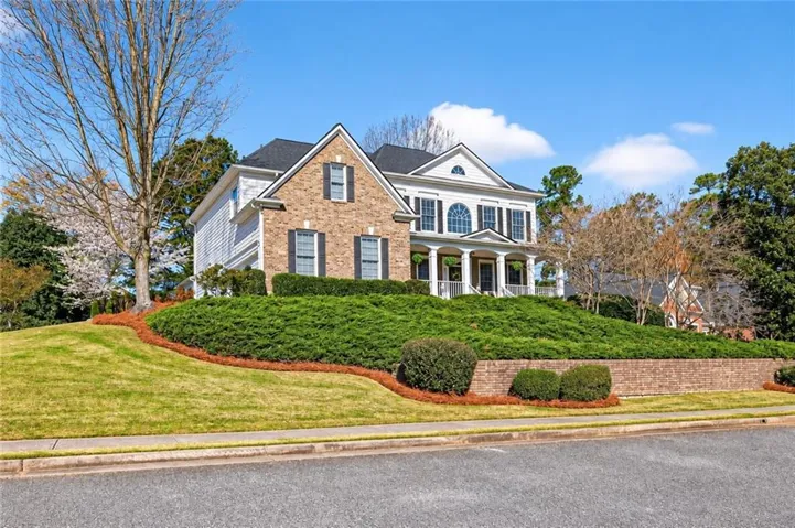 Two-story residence featuring a brick and siding exterior, a front porch with white columns, a dark shingle roof, and a landscaped yard with mature plantings and a retaining wall